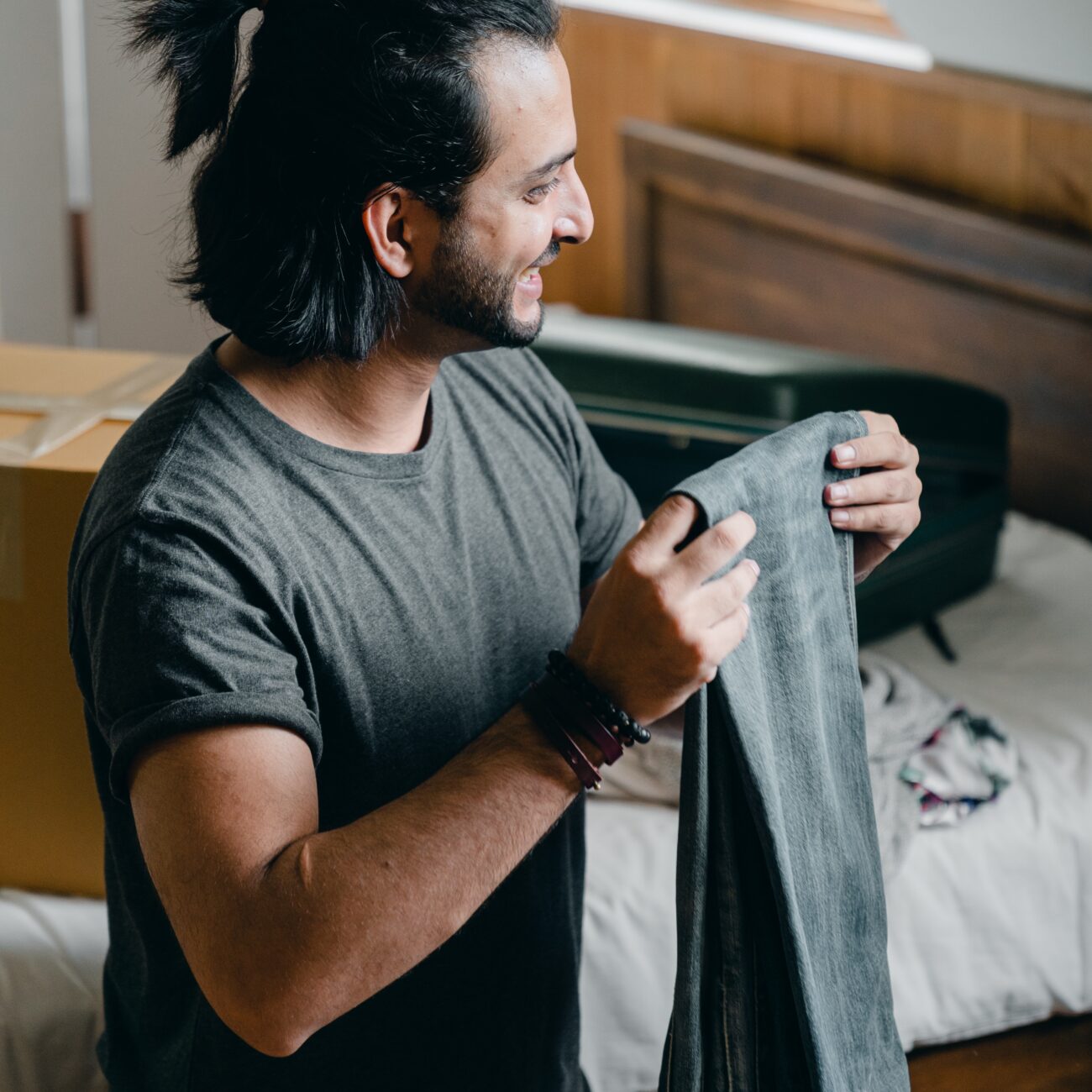 A man smiling while looking at a pair of jeans, while unpacking, in a cozy bedroom setting.