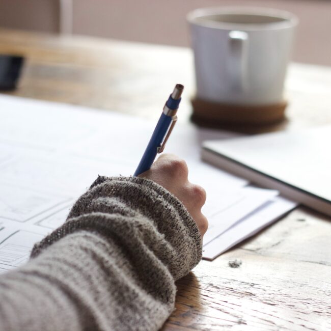 A person writing on a sheet of paper at a wooden table with a steaming cup of coffee nearby, suggesting a moment of creative work or study.