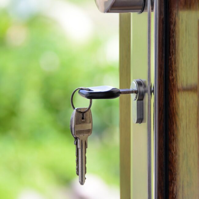 A key inserted into the lock of a wooden door, with a blurry natural backdrop suggesting a tranquil, residential setting.